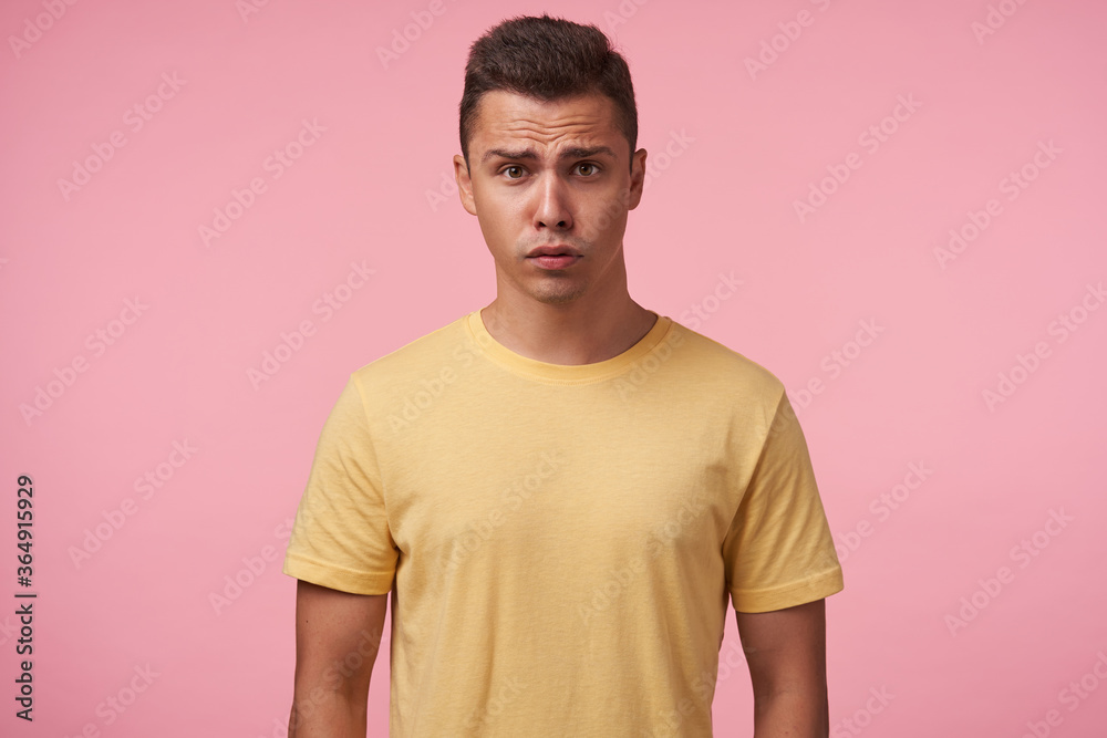Fototapeta premium Bewildered young handsome short haired brunette man grimacing his face while looking confusedly at camera, keeping hands down while posing over pink background