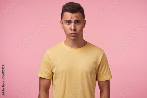 Bewildered young handsome short haired brunette man grimacing his face while looking confusedly at camera, keeping hands down while posing over pink background