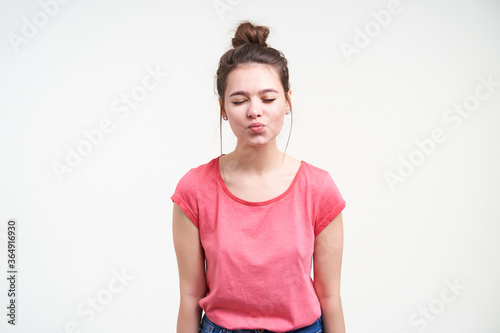 Good looking young charming brown haired woman with natural makeup keeping her eyes closed while folding lips in air kiss, isolated over white background