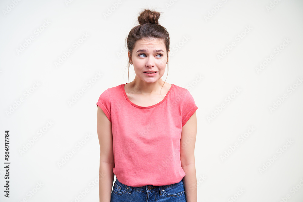 Studio photo of young puzzled brown haired female looking confusedly aside and keeping hands down while standing over white background in pink t-shirt Studio photo of young puzzled brown haired female looking confusedly aside and keeping hands down while standing over white background in pink t-shirt