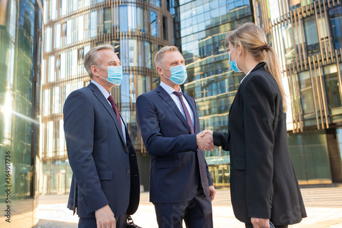 Business men and woman in face masks shaking hands near office buildings, meeting and talking in city. Side view, low angle. Cooperation and coronavirus concept
