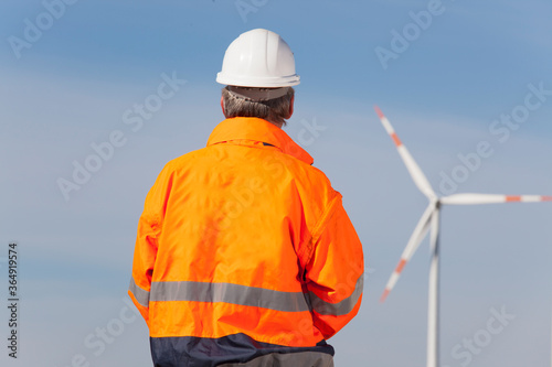 Worker or engineer with hard hat and protecive clothing looking at a windmill of a windfarm