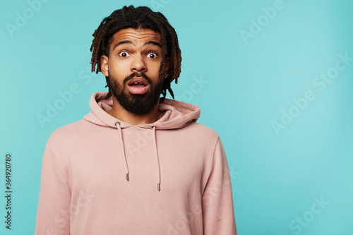 Bemused young pretty brunette bearded man with dark skin looking amazedly at camera with wide eyes and mouth opened while posing against blue background