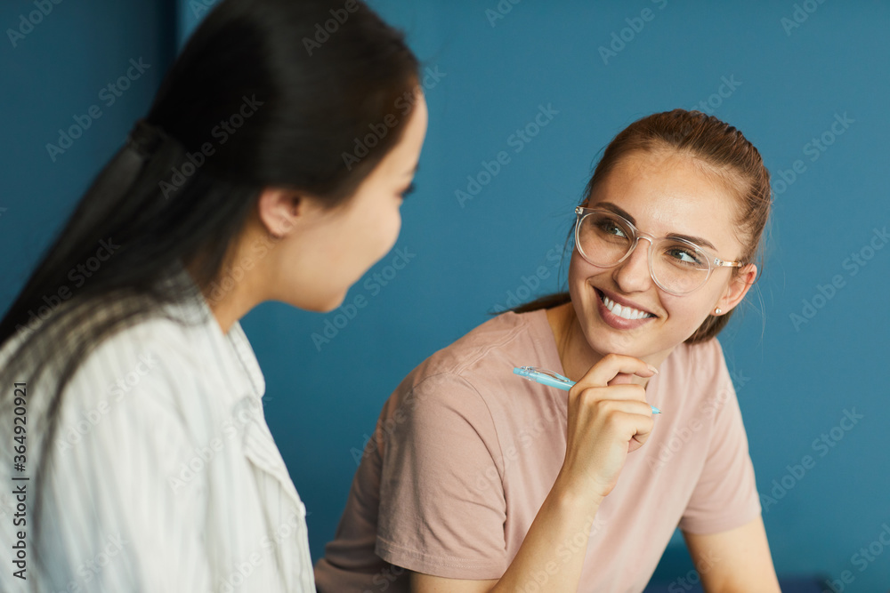 Smiling student girl in eyeglasses holding pen and discussing project ideas with groupmate Smiling student girl in eyeglasses holding pen and discussing project ideas with groupmate