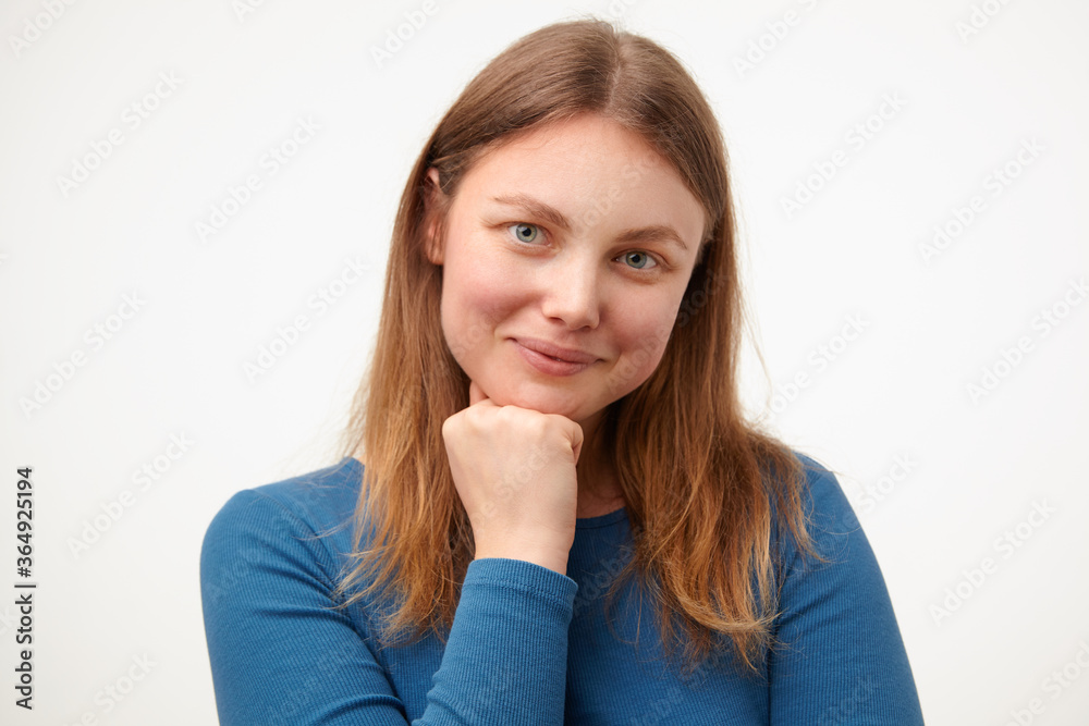 Pleased young female with loose hair leaning chin on raised hand and smiling nicely at camera while posing over white background in casual blue blouse Pleased young female with loose hair leaning chin on raised hand and smiling nicely at camera while posing over white background in casual blue blouse