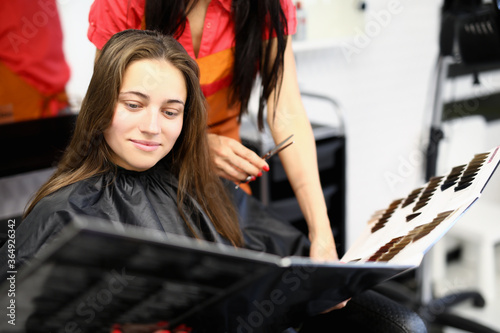 Portrait of cheerful glad female sitting on beautician appointment. Happy young woman holding folder with hair colour samples. Hairdresser and beauty salon concept