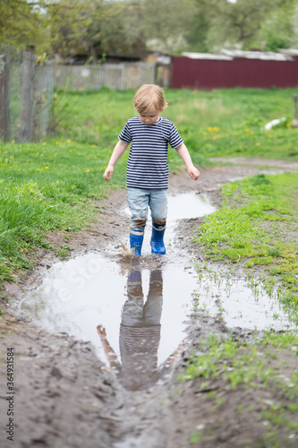a small boy in rubber boots walks through a muddy puddle