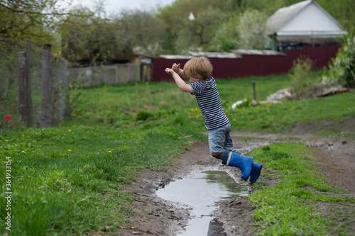 a small boy in rubber boots jumps through a muddy puddle