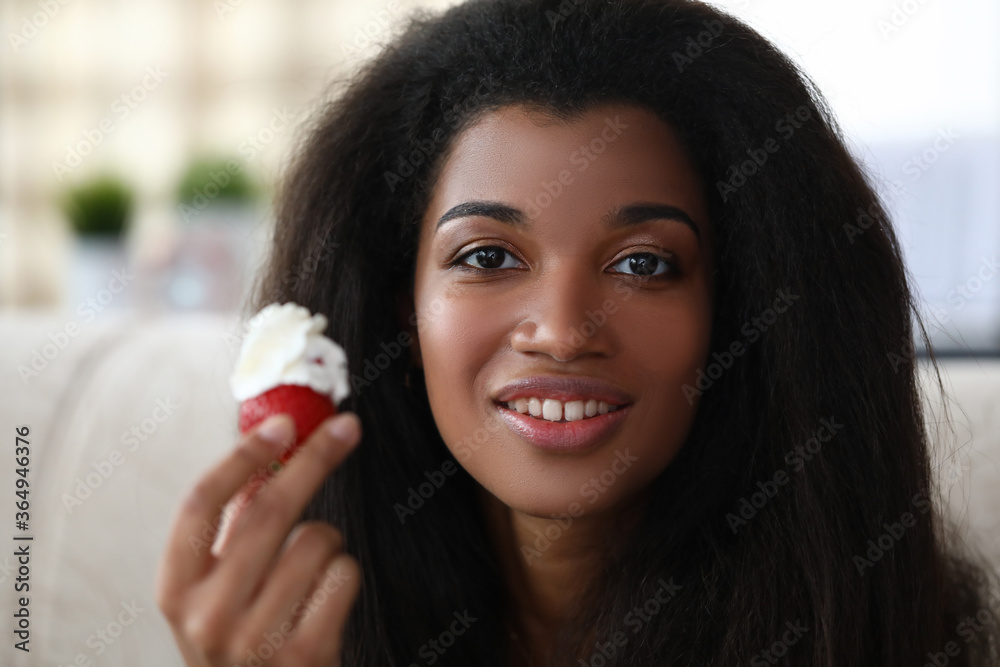 Portrait of happy smiling brunette afro american woman holding mellow strawberry. Cheerful lady posing on camera at home. Relaxation on sofa. Spare time and lunch break concept Portrait of happy smiling brunette afro american woman holding mellow strawberry. Cheerful lady posing on camera at home. Relaxation on sofa. Spare time and lunch break concept