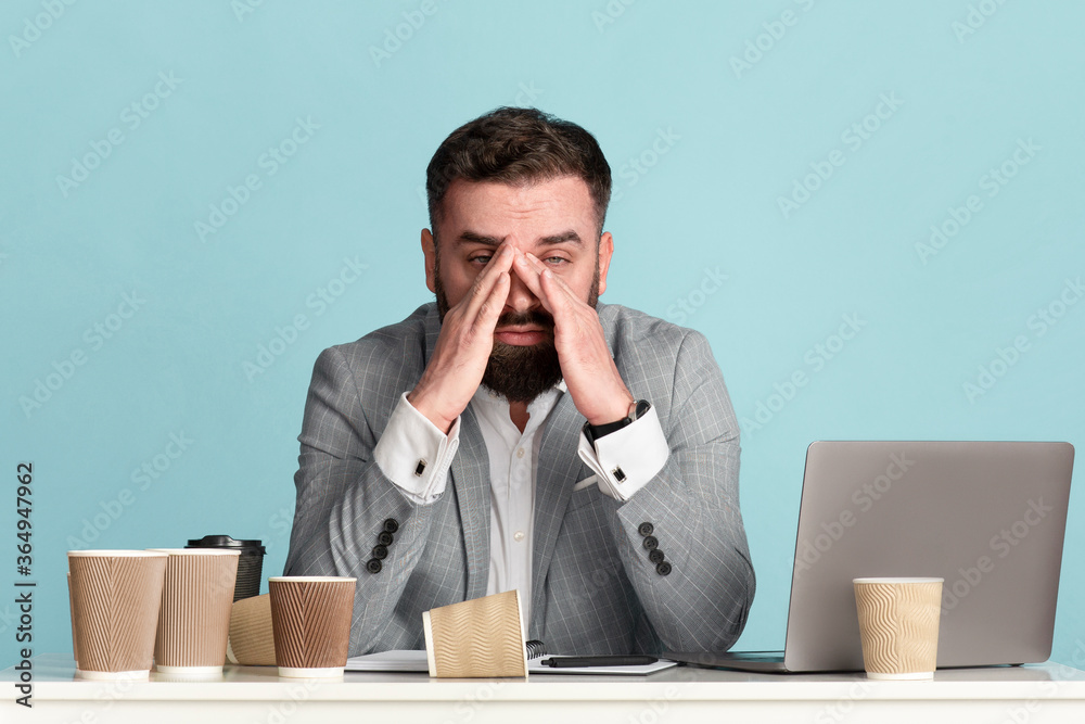 Tired businessman working late at desk with laptop and scattered empty coffee cups, blue background Tired businessman working late at desk with laptop and scattered empty coffee cups, blue background