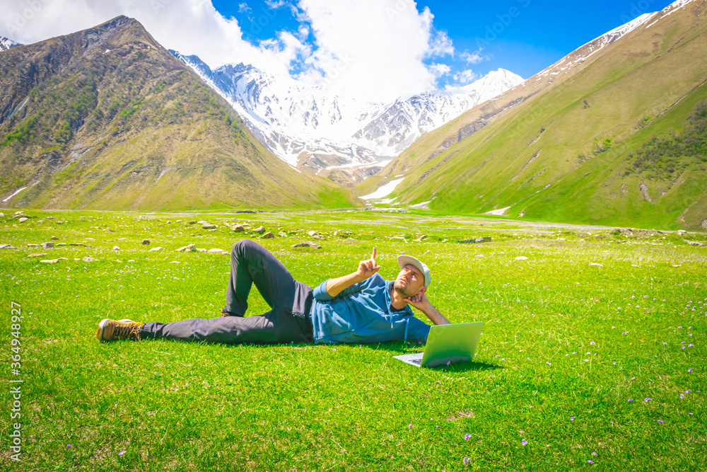Male person in csualclothes lays on the grass and points his finger up with mountains in the background. Inspiration and new ideas born in nature concept. Backgorud image of creativity Male person in csualclothes lays on the grass and points his finger up with mountains in the background. Inspiration and new ideas born in nature concept. Backgorud image of creativity