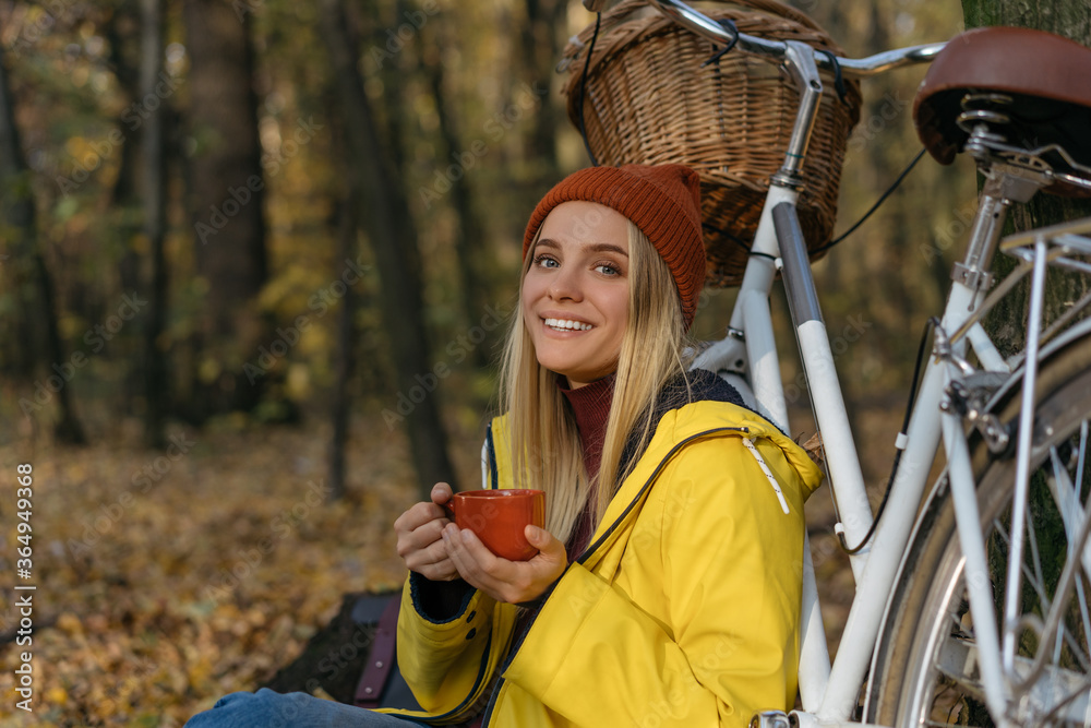 Obraz premium Beautiful woman relaxing in autumn forest, holding cup of coffee, looking at camera , smiling. Active lifestyle concept