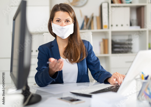 Successful adult business woman in protective medical mask using laptop at workplace