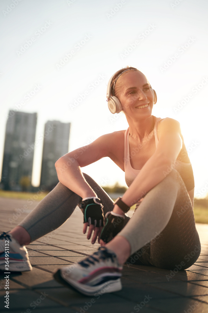 Smiling young fitness woman listening music in headphones and relaxing after jogging or running. Sporty female outdoor