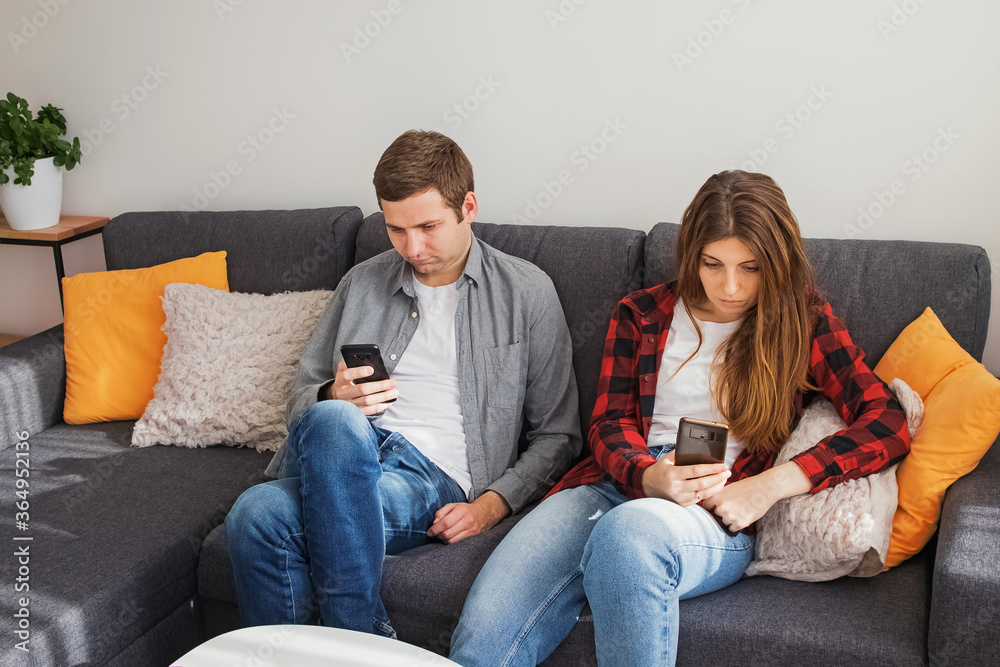 Young couple, man and woman using their mobile phones while sitting on the couch