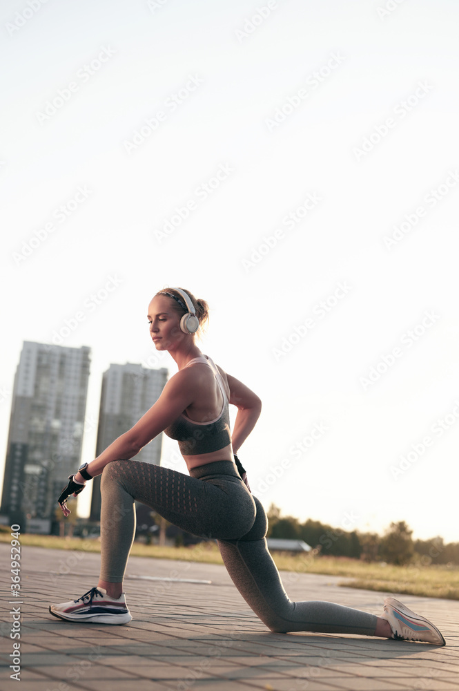 Fototapeta premium Fitness woman taking a break after a workout, strong sporty girl in headphones stretching outdoors, back lit sunset, city view