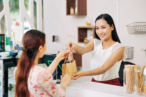 Happy pretty young Vietnamese shop assistant giving paper bag with order to female customer