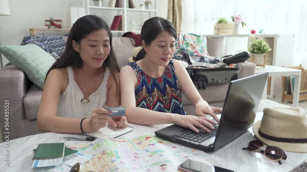 two asian japanese girl friends sitting on floor in house living room searching internet for summer break travel trip. ladies holding credit card buying cheap airplane ticket on internet. e commerce. two asian japanese girl friends sitting on floor in house living room searching internet for summer break travel trip. ladies holding credit card buying cheap airplane ticket on internet. e commerce.