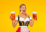 Young sexy Oktoberfest girl waitress, wearing a traditional Bavarian or german dirndl, serving big beer mugs with drink isolated on yellow background. Woman pointing to looking left.