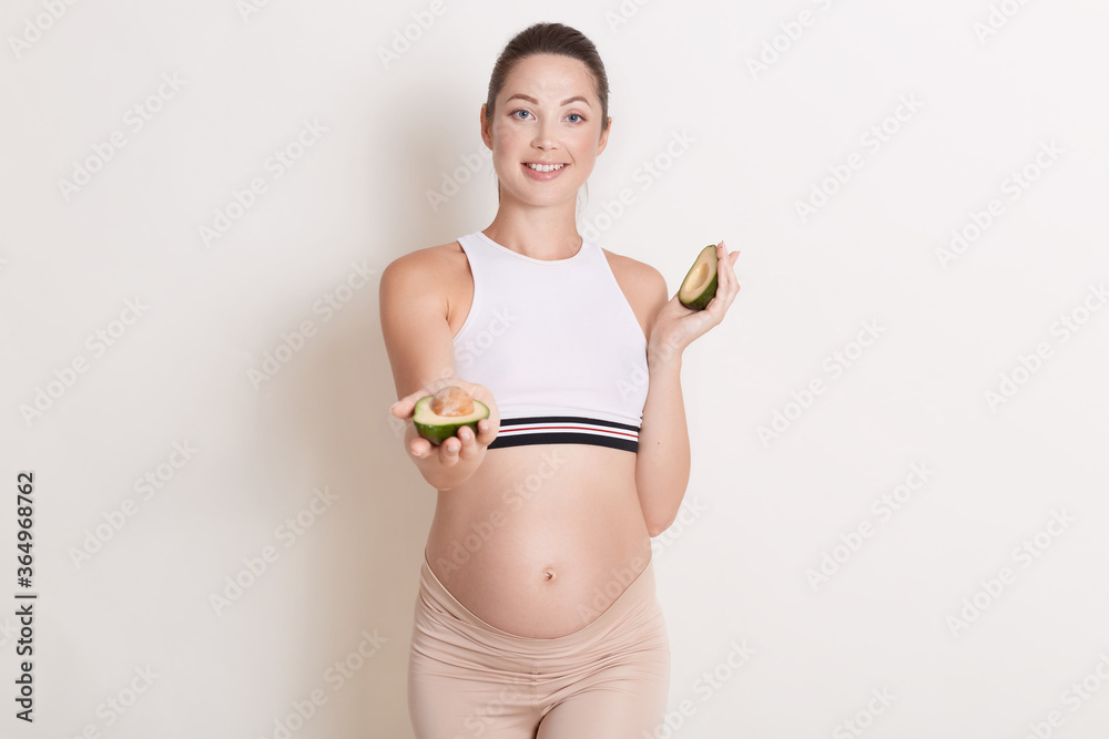 Pregnant woman with avocado in hands, lady awaited baby offers fruit to somebody, standing against white background, girl wearing sporty clothing, has ponytail. Pregnant woman with avocado in hands, lady awaited baby offers fruit to somebody, standing against white background, girl wearing sporty clothing, has ponytail.
