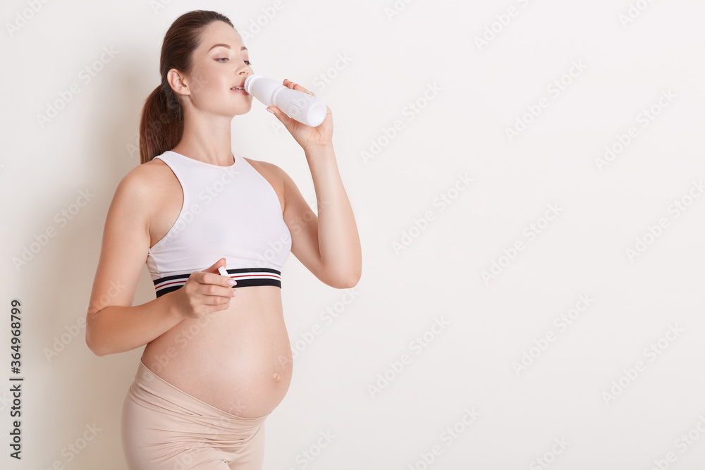 Happy pregnant woman drinking kefir or yogurt from bottle, wearing sporty clothes, showing her bare belly, posing isolated over white background. Copy space. Happy pregnant woman drinking kefir or yogurt from bottle, wearing sporty clothes, showing her bare belly, posing isolated over white background. Copy space.