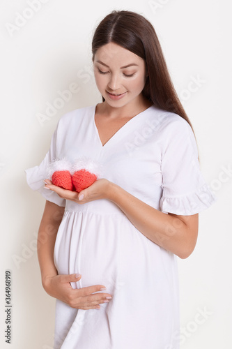 Beautiful pregnant woman wearing white dress. holding in hands small red booties, looking at it with gently smile, lady posing isolated over white background.