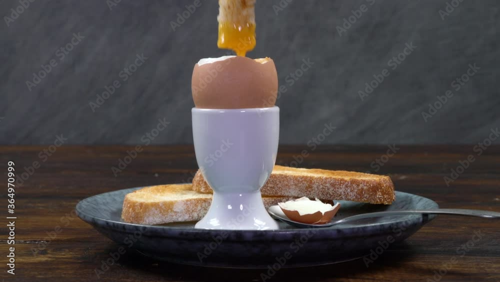 Person dipping a toast soldier into a boiled egg in a white egg cups on a plate with toast and a spoon