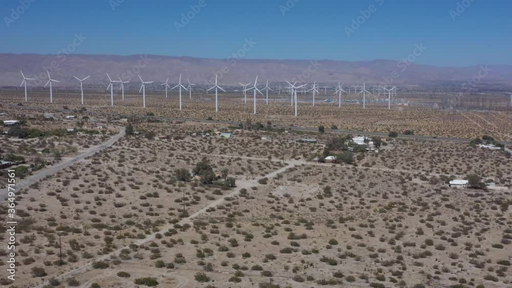 Several renewable wind turbines in countryside arid field with rotating propellers generating energy on bright blue sunny sky day with mountain range in background, aerial static Several renewable wind turbines in countryside arid field with rotating propellers generating energy on bright blue sunny sky day with mountain range in background, aerial static