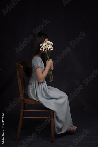Studio portrait of an anonymous young girl in long blue dress sitting on a chair with bouquet of  white with purple arums in front of her face photo