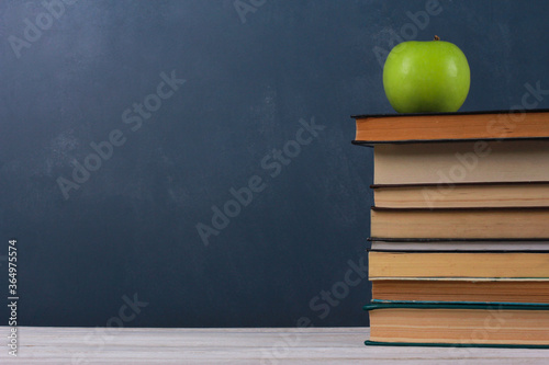 Green apple on big stack of books,  on white wooden desk with blackboard and chalk streaks in background. Side view, copy space, close-up. Learning, education, knowledge, library, love reading concept