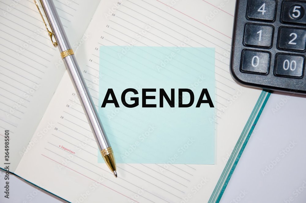 The word Agenda is written on a paper blue block near a calculator and notepad. pen next The word Agenda is written on a paper blue block near a calculator and notepad. pen next