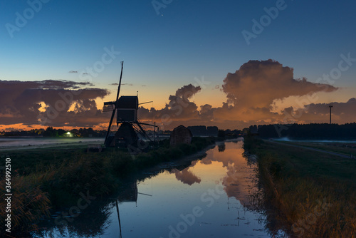 Night image of a Dutch windmill along the water of a canal. Layer of fog in the fields and dramatic clouds in the background.