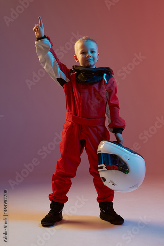 Fotografie, Obraz Boy child racer with helmet and his hand up, standing in neon light