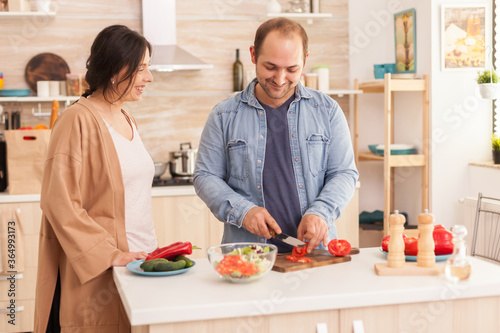 Man slicing tomatoes for salad on cutting board in kitchen while wife is smiling. Happy in love cheerful and carefree couple helping each other to prepare meal