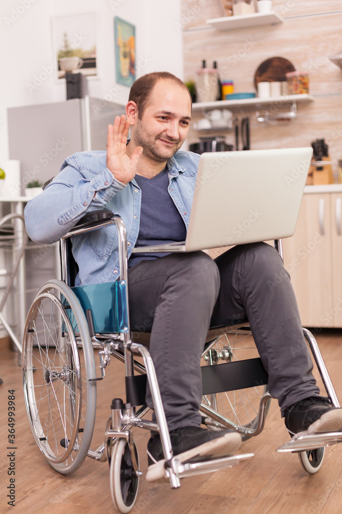 Businessman in wheelchair waving during a video conference on laptop in kitchen while is preparing food. Disabled paralyzed handicapped man with walking disability integrating after an accident. Businessman in wheelchair waving during a video conference on laptop in kitchen while is preparing food. Disabled paralyzed handicapped man with walking disability integrating after an accident.