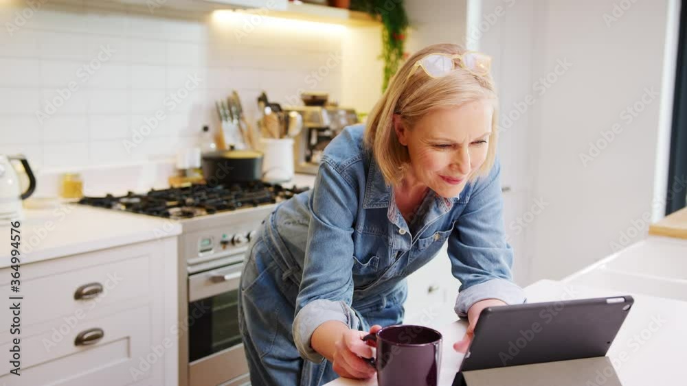 Mature woman at home in kitchen drinking coffee and looking at digital tablet - shot in slow motion Mature woman at home in kitchen drinking coffee and looking at digital tablet - shot in slow motion