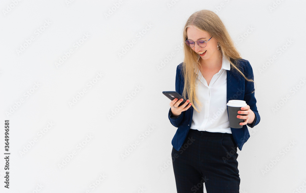 A young caucasian blonde business woman is standing in front of a white wall, smiling, looking at the phone and holding coffee in her hand.  Concept of successful young women. Lifestyle concept. 