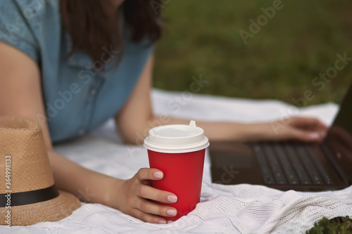 Young woman lies on a plaid and using laptop in the park. Working on laptop outdoors.