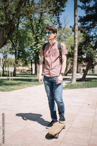 Cool guy with sunglasses posing with skateboard. Portrait of man standing in park with skateboard during sunny day.