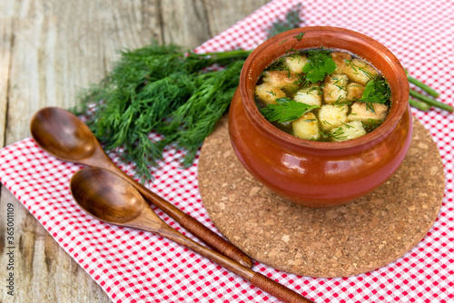 Delicious soup with croutons, herbs in a clay pot on a red checkered napkin. Pot with broth and dill in the village.