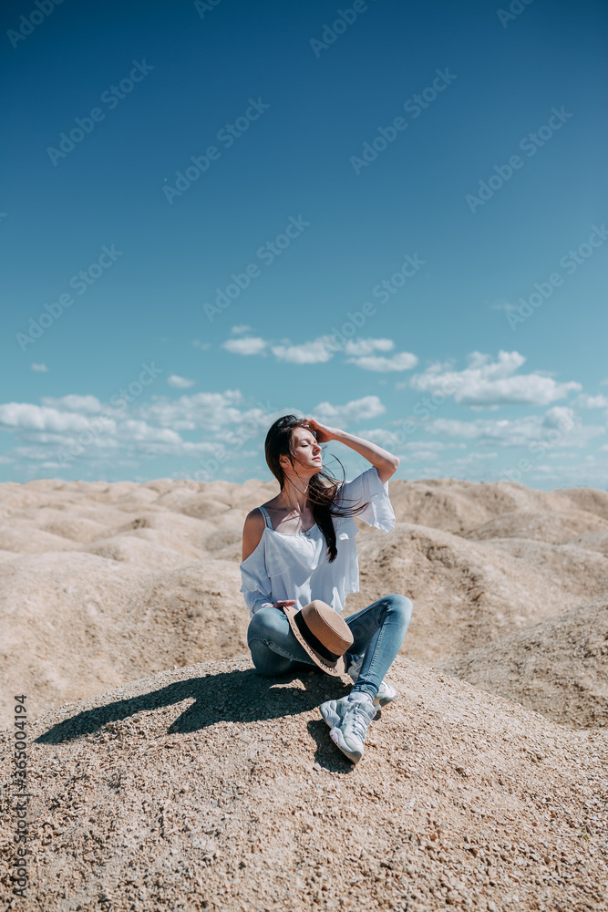 Woman in blue jeans, white shirt and a hat seatting on rocks. White hills of refuse pile and blue sky. Woman in a desert on spoil heap.