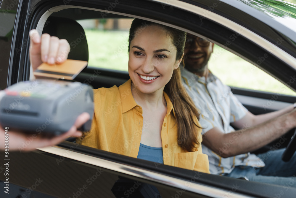 Selective focus of smiling woman paying with credit card near husband in car and worker of gas station holding payment terminal