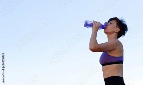 A girl with short hair in a purple top and dark pants drinks water from a bottle when doing sports. Against the sky.