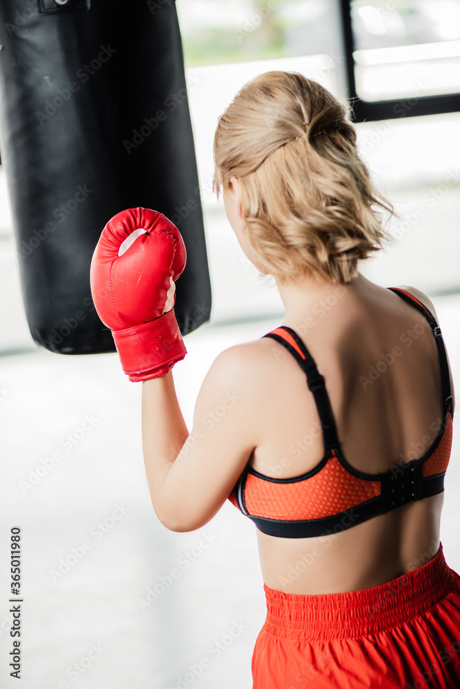 back view of sportive girl in boxing gloves exercising with punching bag