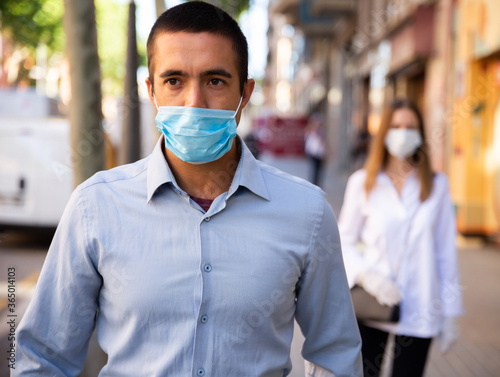 Focused adult man wearing medical mask walking along city street in sunny spring morning. New life reality during coronavirus pandemic.