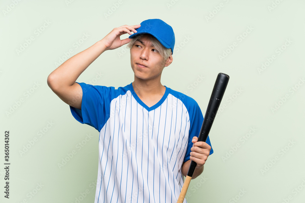 Young asian man playing baseball over isolated green background having doubts and with confuse face expression