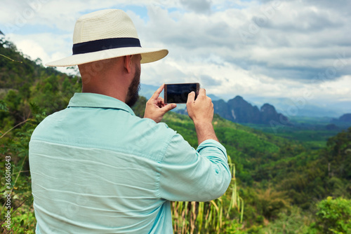 Back view of a young man traveler is taking photo with cell telephone camera of a beautiful jungle landscape. Young male wanderer is shooting video on mobile phone during summer adventure in Asia