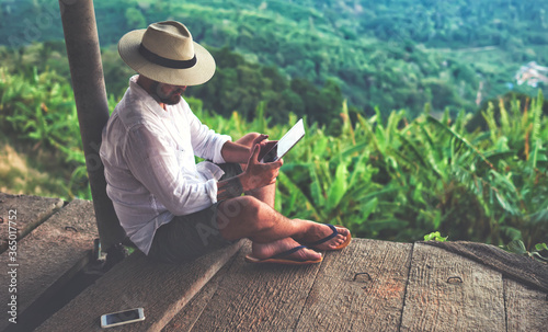 Man traveler is using digital tablet, while is sitting against beautiful Asian scenery during summer journey. Male wanderer is holding touch pad, while is relaxing outdoors during his trip in Thailand photo