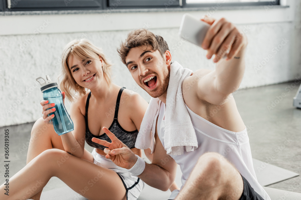 selective focus of cheerful man showing peace sign and taking selfie with girl selective focus of cheerful man showing peace sign and taking selfie with girl