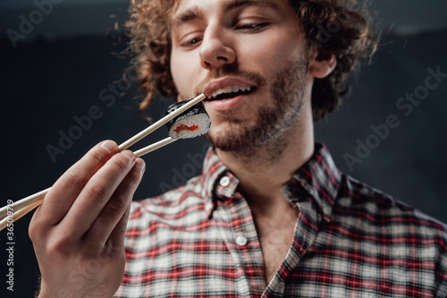 Young stylish man with curly hairstyle wearing checkered shirt eating sushi rolls on the dark background